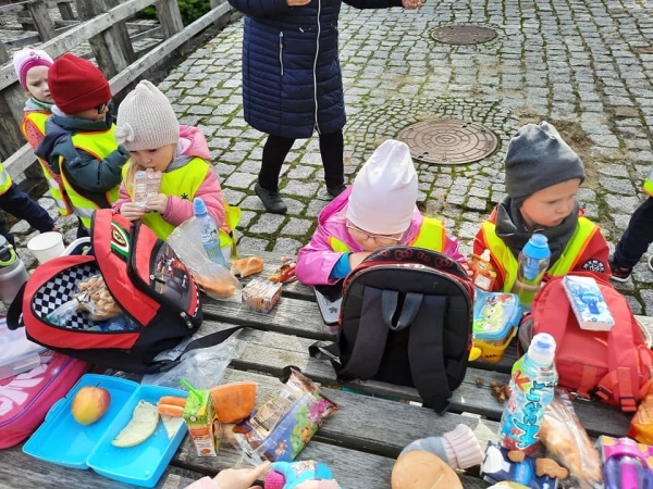 Dzieci w kurtkach i w czapkach siedzą przy 
drewnianym stole 
i jedzą przekąski.